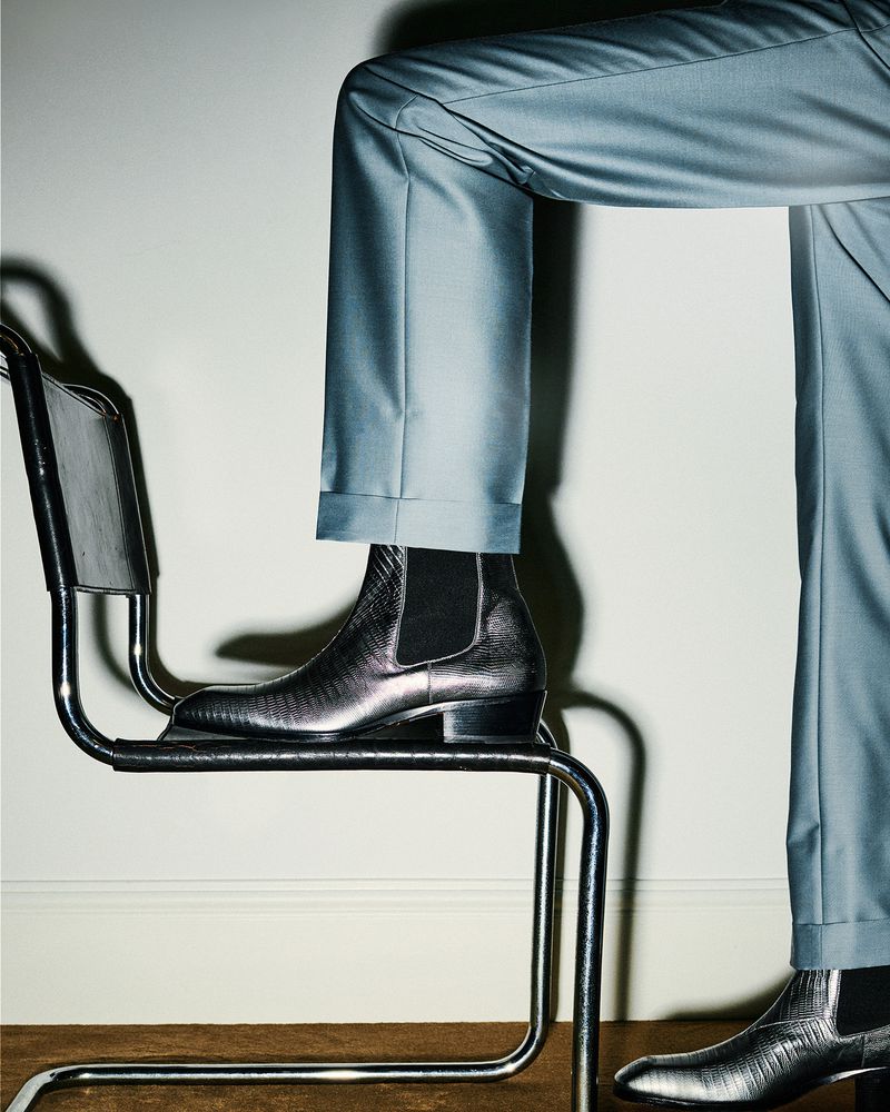 Lower portion of a person in a light blue suit and silver metallic boots standing on a chrome chair against a light background.