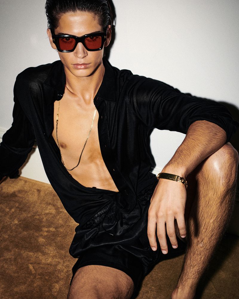 A tanned male model poses shirtless with a black shirt and shorts, red sunglasses, and a gold chain necklace in a studio shot.