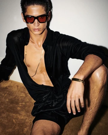 A tanned male model poses shirtless with a black shirt and shorts, red sunglasses, and a gold chain necklace in a studio shot.