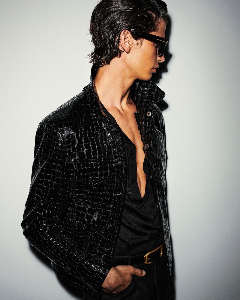 Man in black crocodile jacket and sunglasses poses in a fashion studio. He has wet hair and a gold belt buckle.