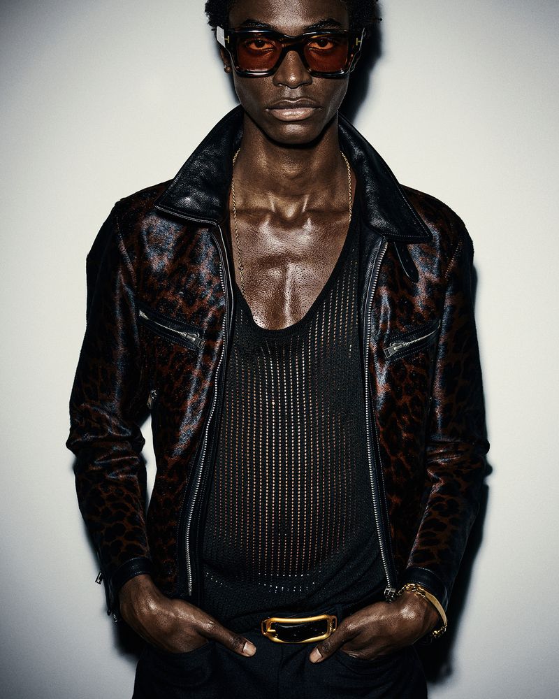 Portrait of a Black male model with curly hair wearing sunglasses, a black mesh top, a jacket, and a gold belt, posing in a studio.