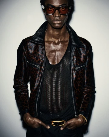 Portrait of a Black male model with curly hair wearing sunglasses, a black mesh top, a jacket, and a gold belt, posing in a studio.