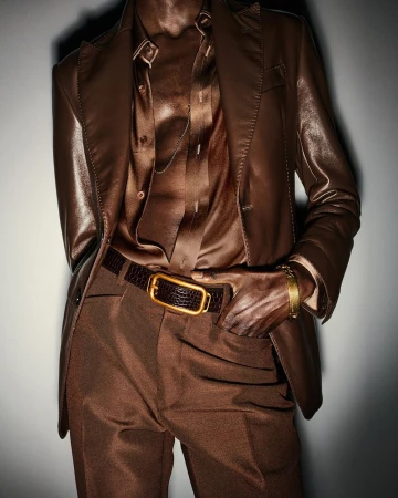 A person wears a brown leather suit with a snakeskin belt and a gold bracelet against a plain backdrop in a fashion-forward studio shot.