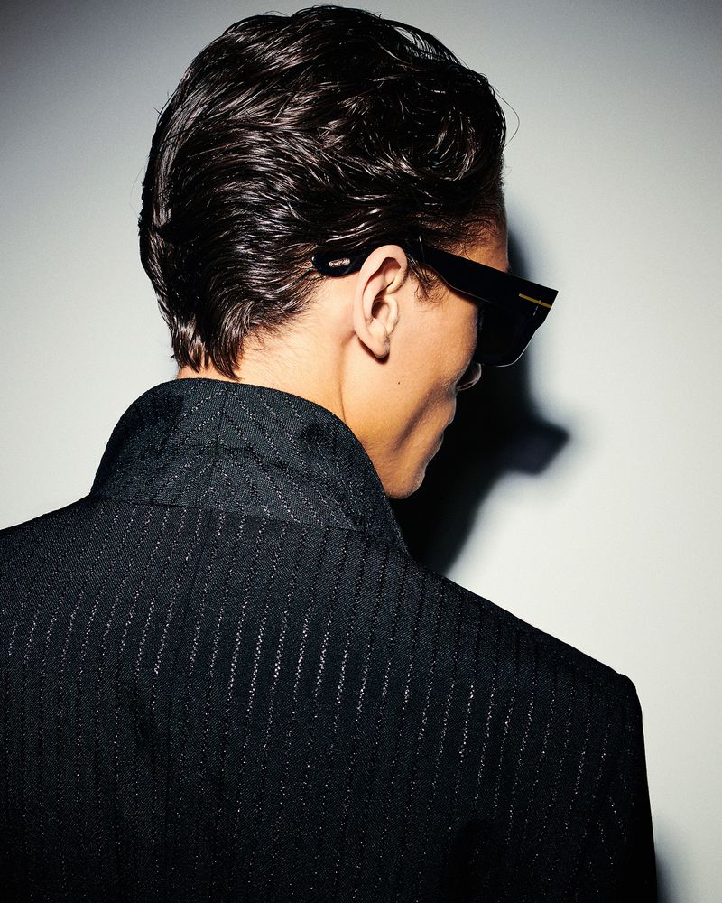 An adult male with dark, slicked-back hair wears sunglasses and a black suit, viewed from the back against a neutral studio backdrop.
