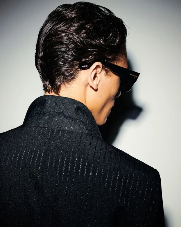 An adult male with dark, slicked-back hair wears sunglasses and a black suit, viewed from the back against a neutral studio backdrop.