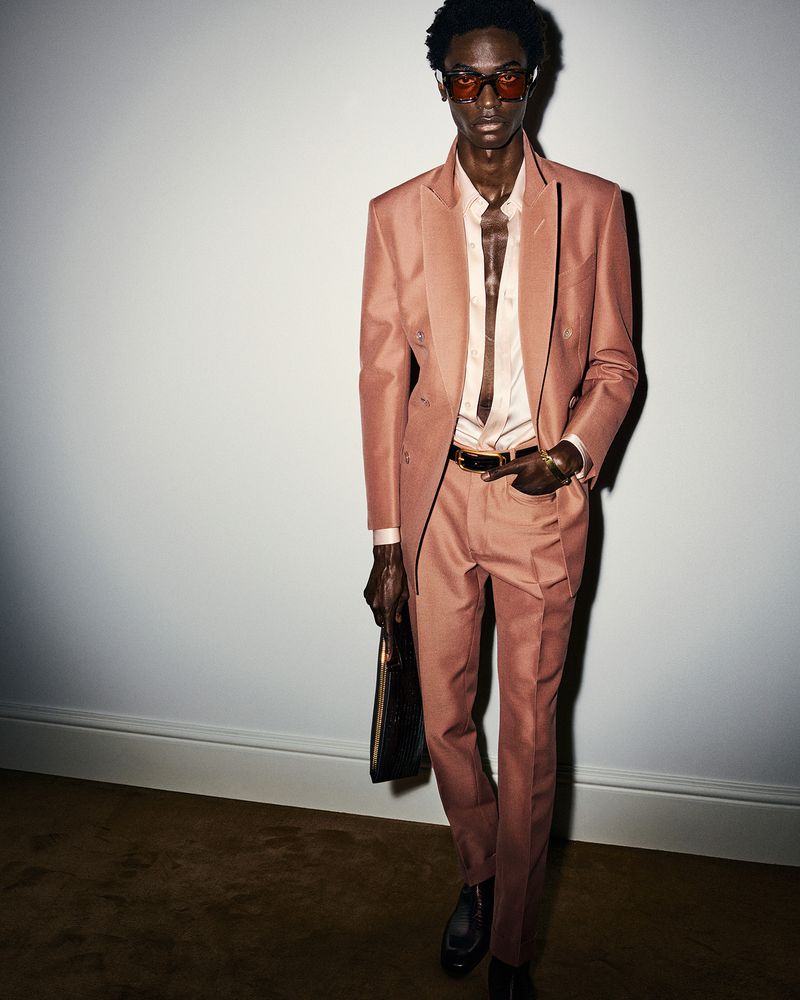 An adult male model with an open-collar shirt and sunglasses poses in a salmon-colored suit against a plain white wall backdrop.