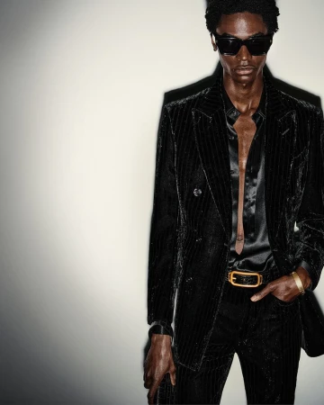 Portrait of a Black male fashion model wearing a black pin-striped suit, black shirt, sunglasses, and gold belt, posing against a light wall.
