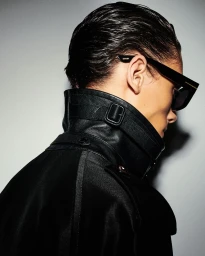 Profile view of a man wearing black sunglasses and a high-collared black leather jacket against a light grey studio background.