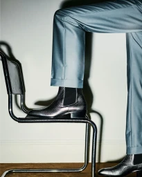 Model wearing metallic chelsea boots and light blue trousers resting a foot on a metal framed chair