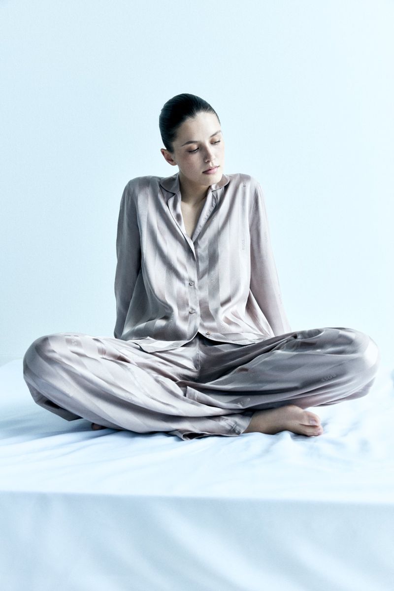 Adult woman in a relaxed cross-legged pose, wearing a satin loungewear set against a soft light blue backdrop in a minimal indoor setting.