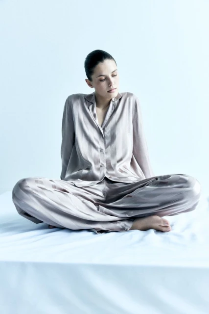 Adult woman in a relaxed cross-legged pose, wearing a satin loungewear set against a soft light blue backdrop in a minimal indoor setting.
