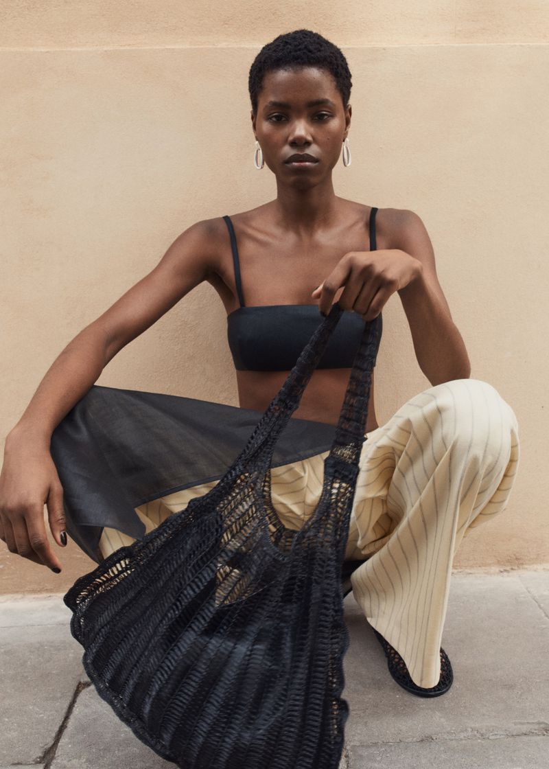 Fashionable African-American woman in a black top and skirt poses with a black mesh tote bag against a tan background.