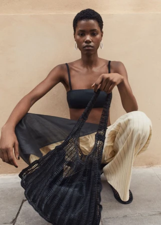 Fashionable African-American woman in a black top and skirt poses with a black mesh tote bag against a tan background.