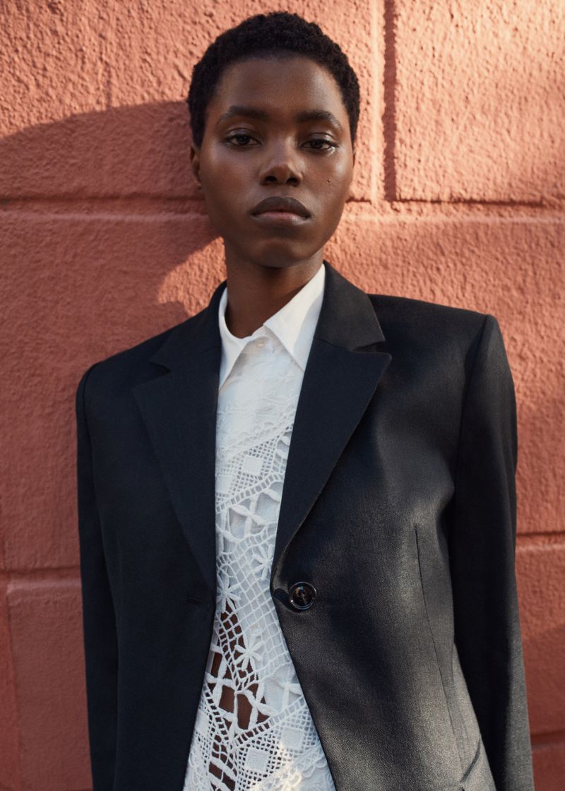 Woman in a black blazer and white lace top poses in front of a textured red brick wall in a medium shot with natural lighting.