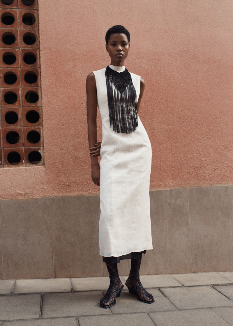 Woman in a white midi dress with black fringe detail stands against a textured peach wall outdoors in a fashion editorial shot.