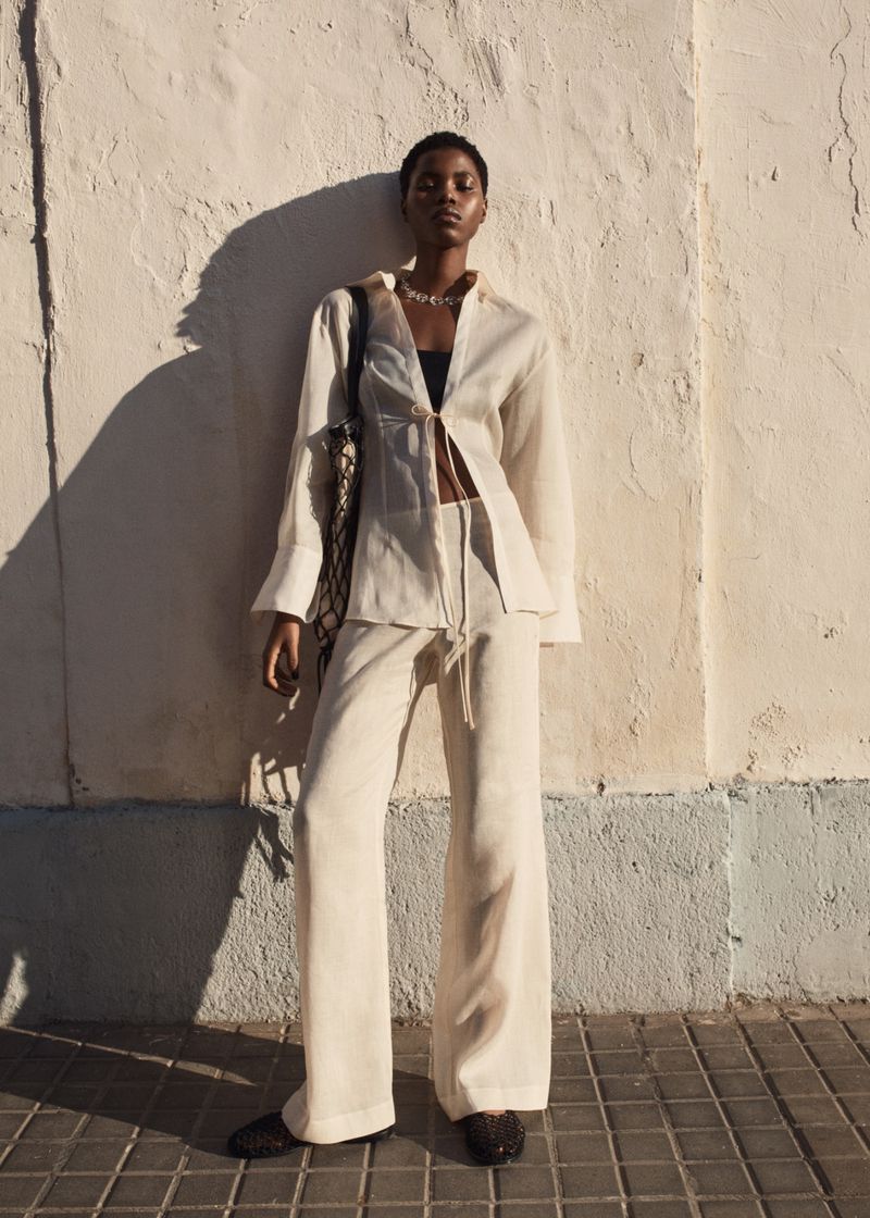An adult woman in a linen suit stands against a wall, carrying a shoulder bag, bathed in warm light with soft shadows.