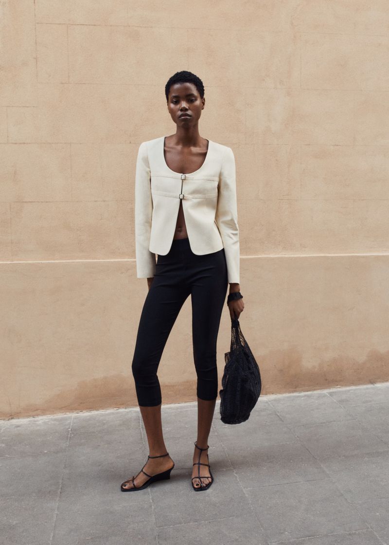 A slender black woman models fashion outdoors, wearing a cream jacket, black pants, and strappy sandals against a neutral wall background.