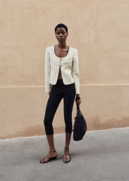 Model wearing a cream jacket and black trousers standing against a textured wall holding a mesh bag