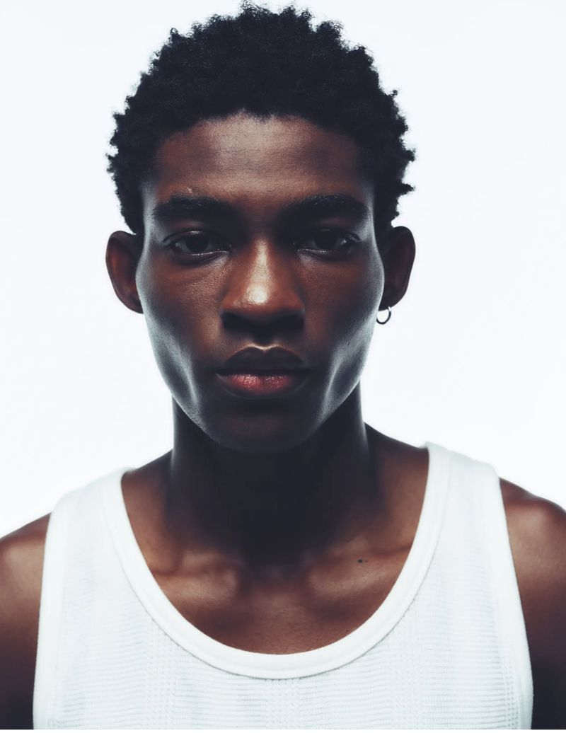 Portrait of a young African American man with dark curly hair, wearing a white tank top, posing against a bright white background.
