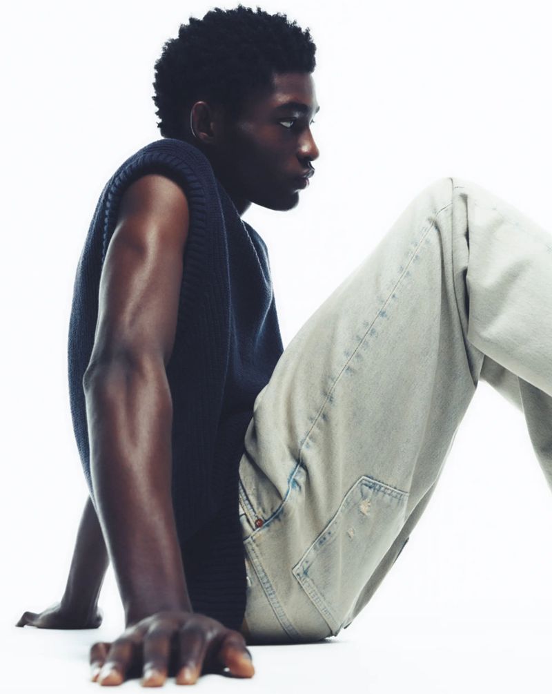 A dark-skinned man in a sleeveless knit top and light-wash jeans poses against a white backdrop in a close-up fashion shot.