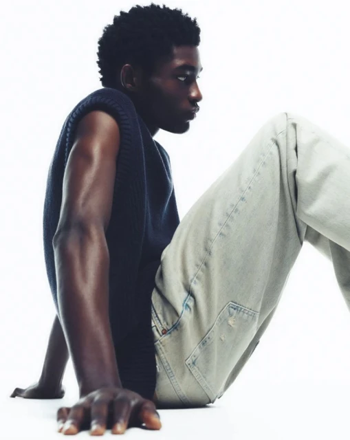 A dark-skinned man in a sleeveless knit top and light-wash jeans poses against a white backdrop in a close-up fashion shot.