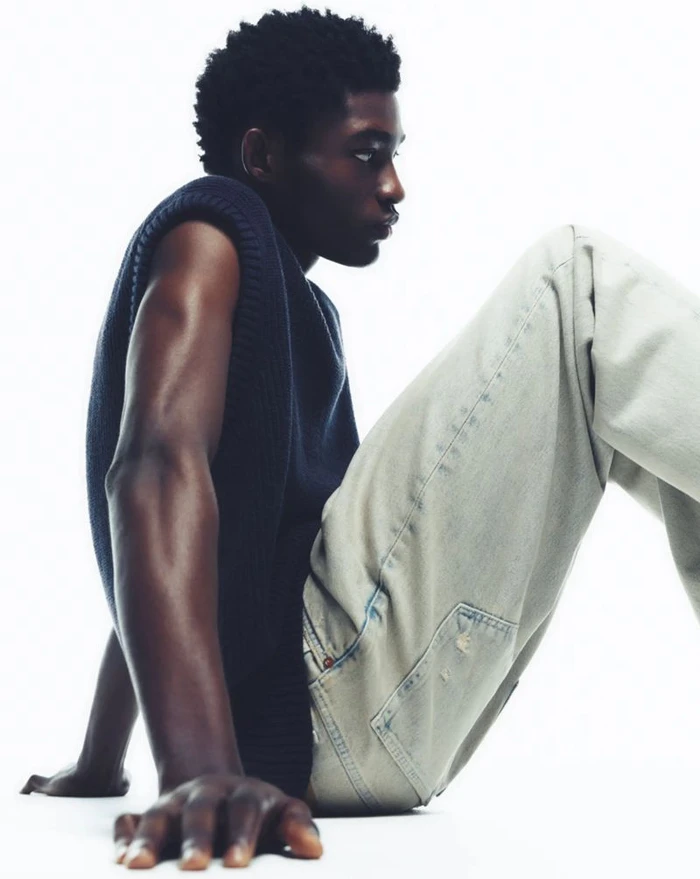 Black man wearing a dark sleeveless sweater and light jeans seated against a white background for a Samsoe SS24 lookbook portrait