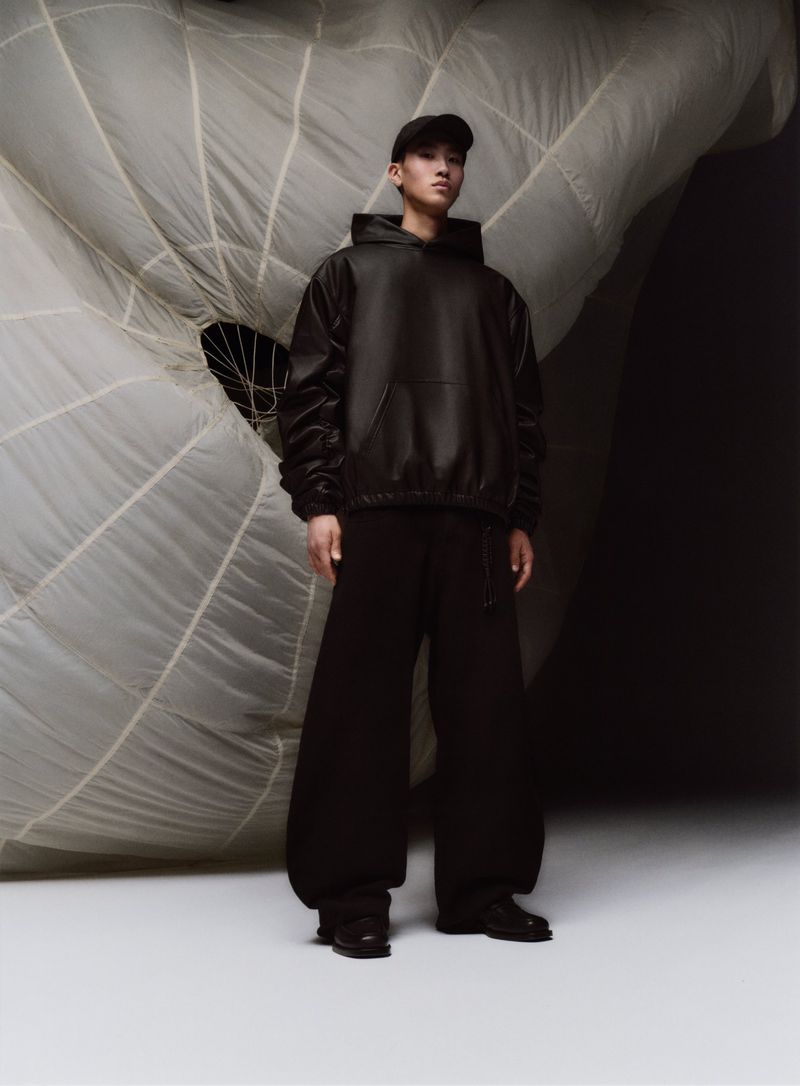 An Asian adult male in a black hoodie and sweatpants stands in a photo studio against a draped white parachute backdrop for fashion editorial.