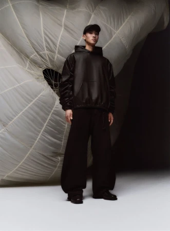 An Asian adult male in a black hoodie and sweatpants stands in a photo studio against a draped white parachute backdrop for fashion editorial.