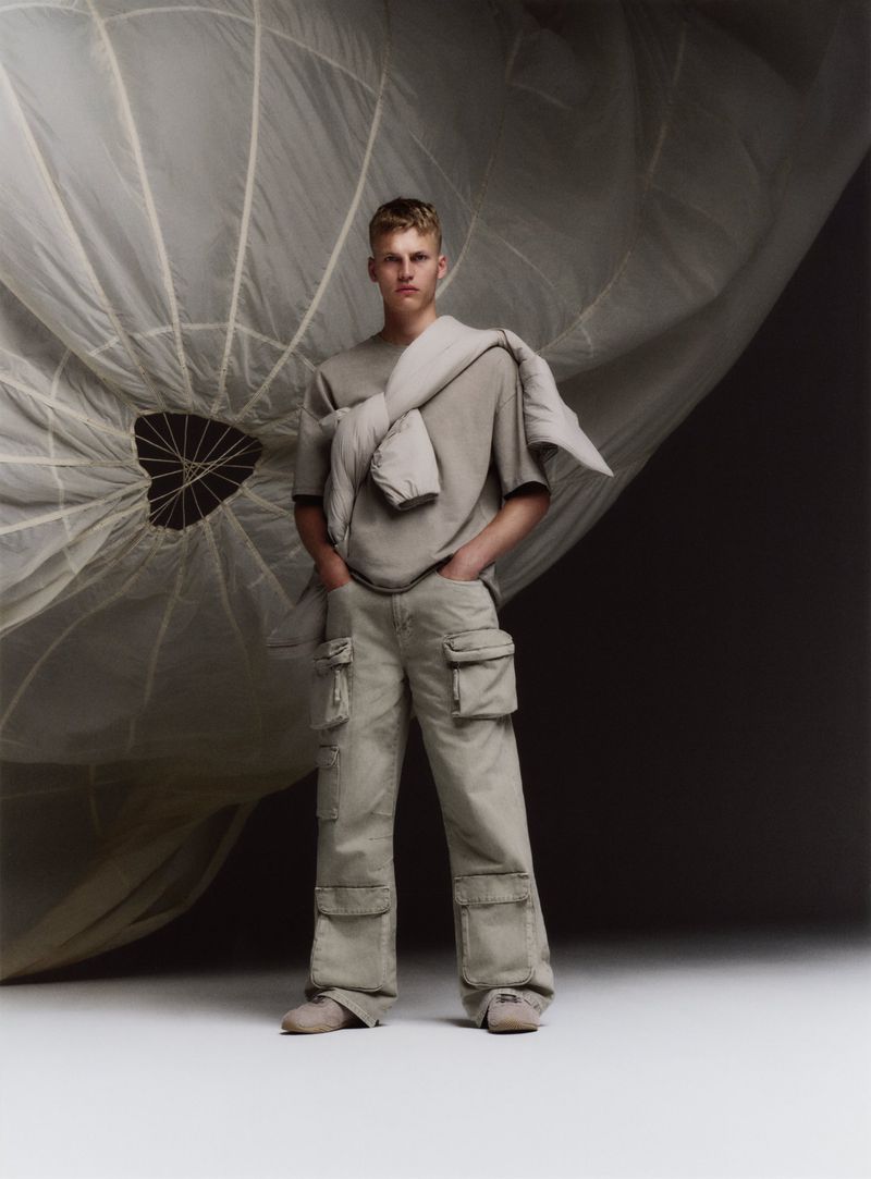 Fashion shot of a young adult man in neutral casualwear, posed with hands in pockets against a large parachute, in a studio setting.