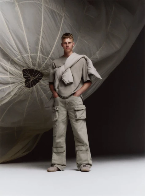 Fashion shot of a young adult man in neutral casualwear, posed with hands in pockets against a large parachute, in a studio setting.