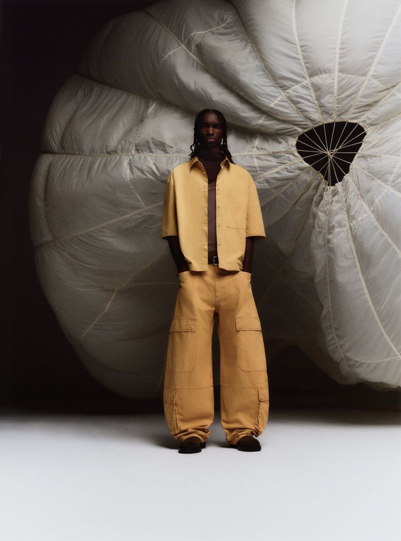A man stands in front of a large parachute. He's wearing tan cargo pants and a matching shirt. The background is a dark studio.