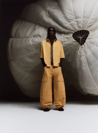 A man stands in front of a large parachute. He's wearing tan cargo pants and a matching shirt. The background is a dark studio.