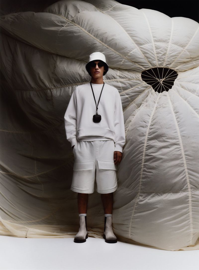 Man posing in white shorts and sweatshirt against a parachute, wearing sunglasses, hat and boots for a fashion advertisement.