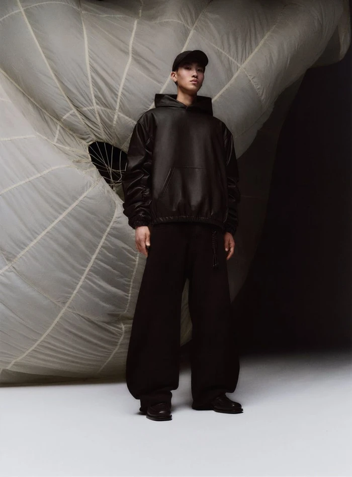 Model in black hoodie and cap standing against a large white draped fabric backdrop for Zara Basez