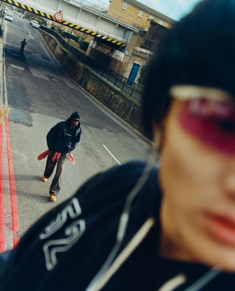 Elevated eye-level shot of two adult males on an asphalt road under a yellow-marked bridge, one figure more prominent and blurred.