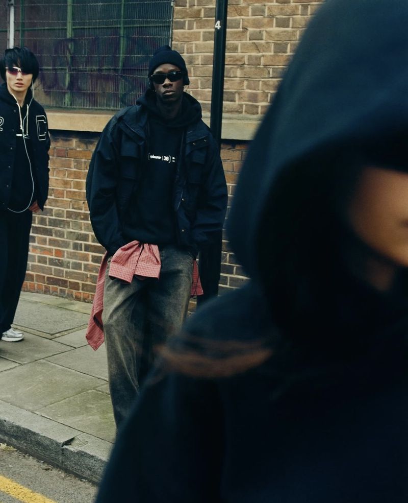 Three people stand in an urban environment. One person is in focus, wearing a dark hoodie, while the other two are out of focus.