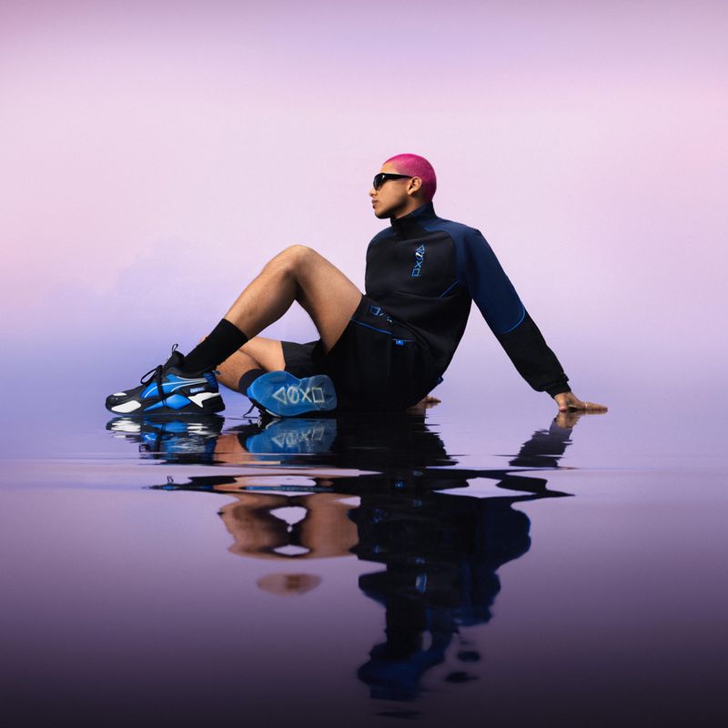 Male model with pink hair poses in athletic wear near water reflecting a pink and purple sky, showcasing sneakers with blue accents.