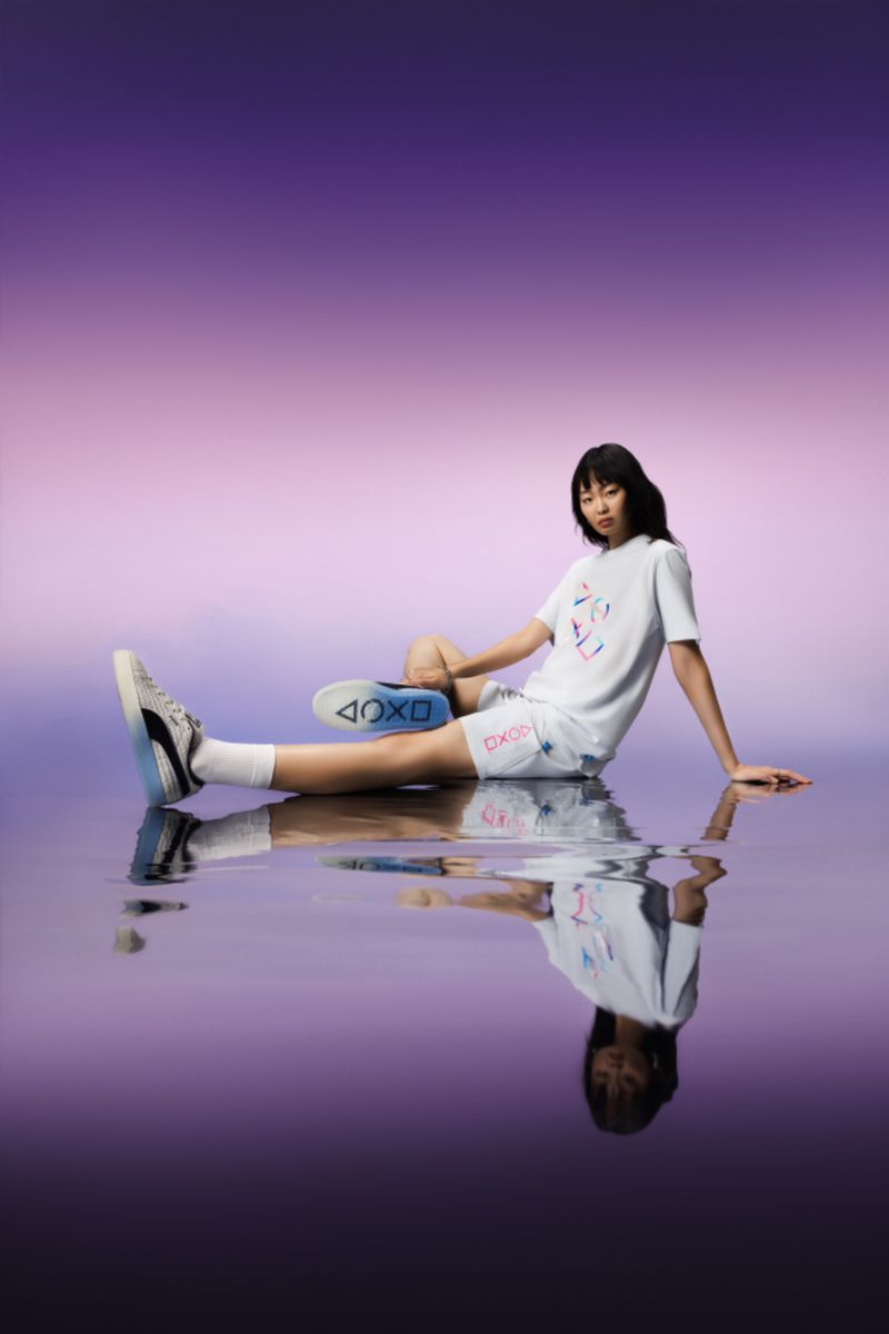 A young woman poses indoors wearing Puma sneakers and a white Playstation t-shirt and shorts against a gradient purple background.