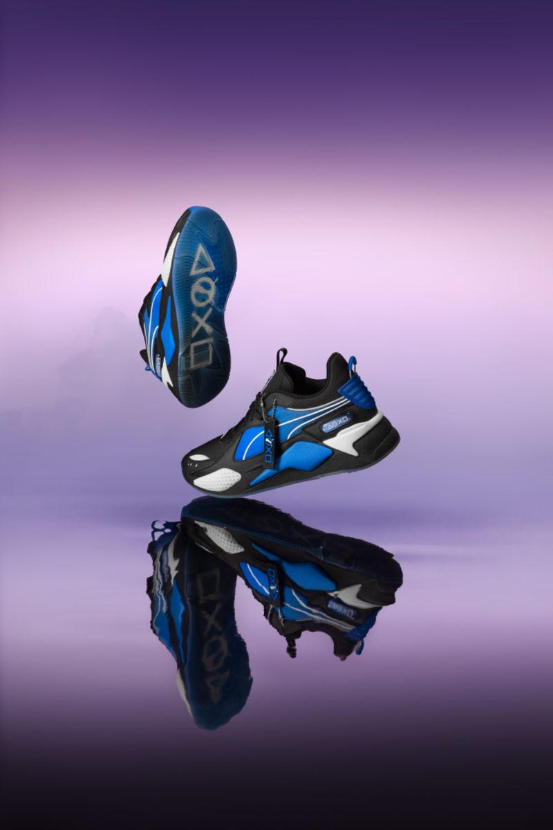 Black and blue Puma RS-X sneakers float in a studio shot against a purple background with their reflection showing on a surface.