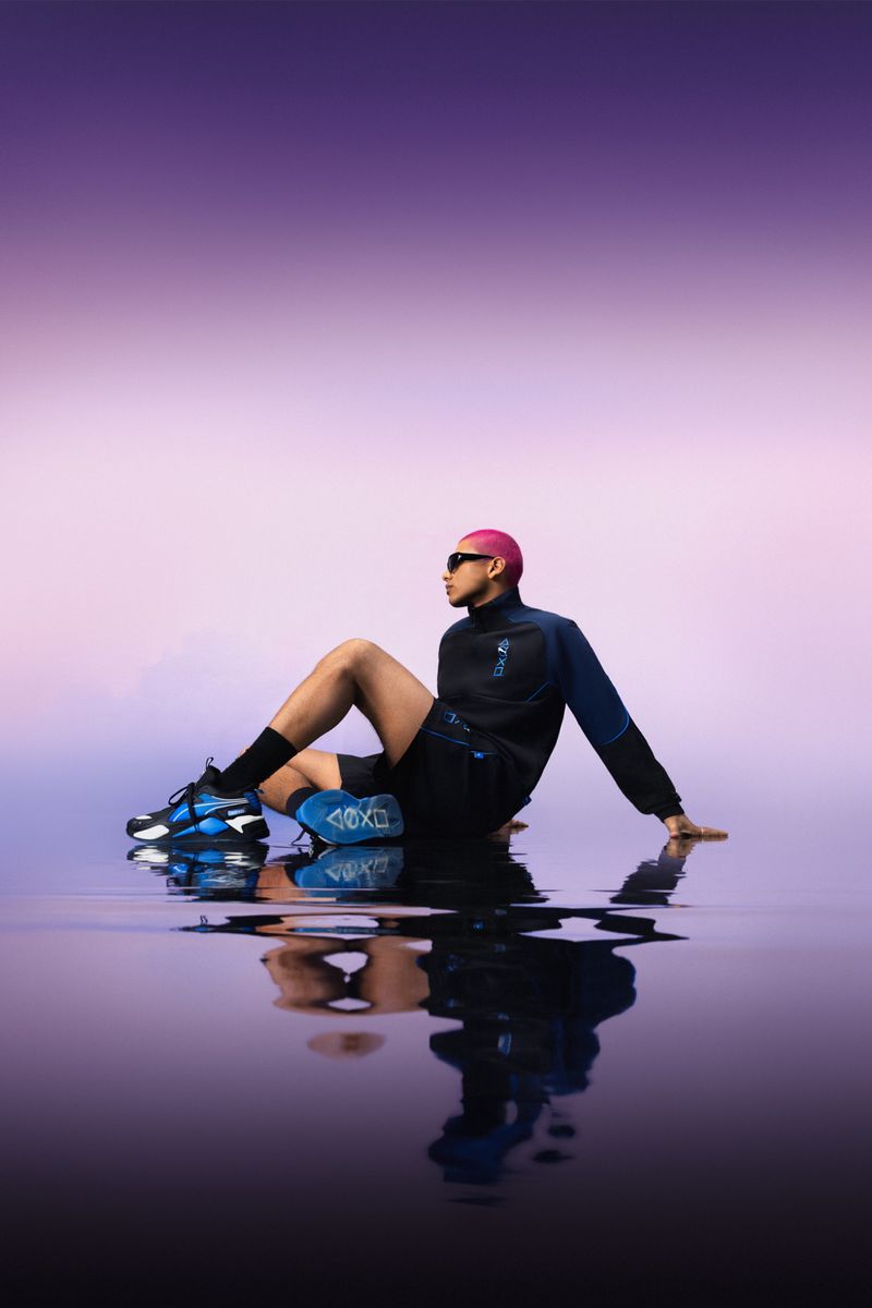 Man with pink hair and sunglasses poses in a wet surface wearing a black tracksuit and sneakers with a purple and pink gradient background.