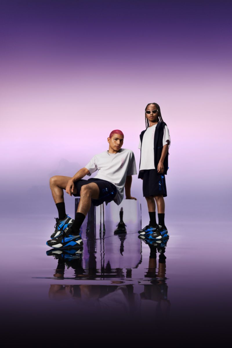 Two young adult models, one seated on a clear cube, showcase black and blue athletic shoes against a graduated purple backdrop in a studio shot.