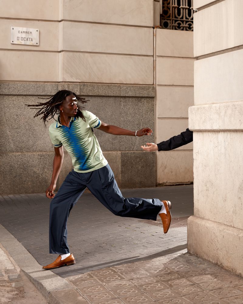 An adult male runs on a city street wearing a polo shirt and dress pants, with another person's hand reaching out toward him.