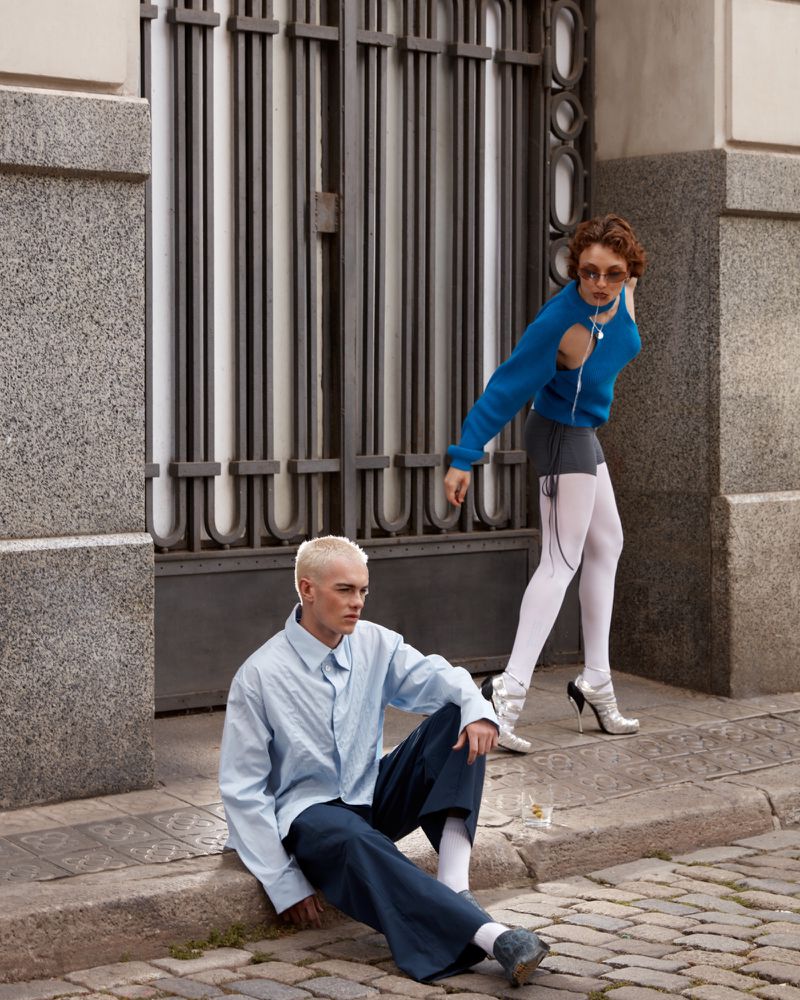Two fashion models pose in an urban setting: one seated wearing a blue shirt and one standing in a blue sweater and sunglasses.