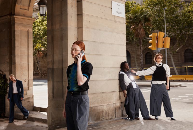 Four people in a street setting; one woman uses a cell phone, two people lean on a wall, and one person walks under an archway.