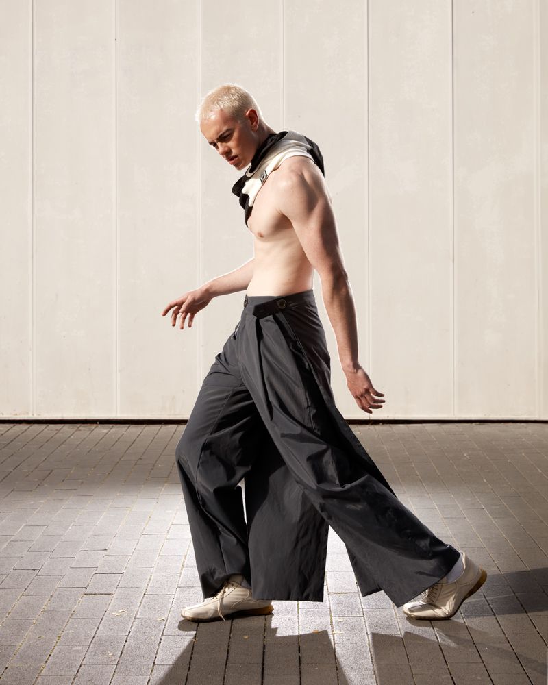 Male fashion model with bare chest wearing wide leg trousers in a studio setting with a minimalistic background and indoor lighting.