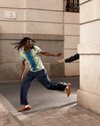 Man with dreadlocks in a patterned polo shirt and dark trousers reaching out near a stone building wall