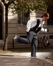 Red-haired model in a white shirt and dark trousers stepping forward outdoors with a leather handbag