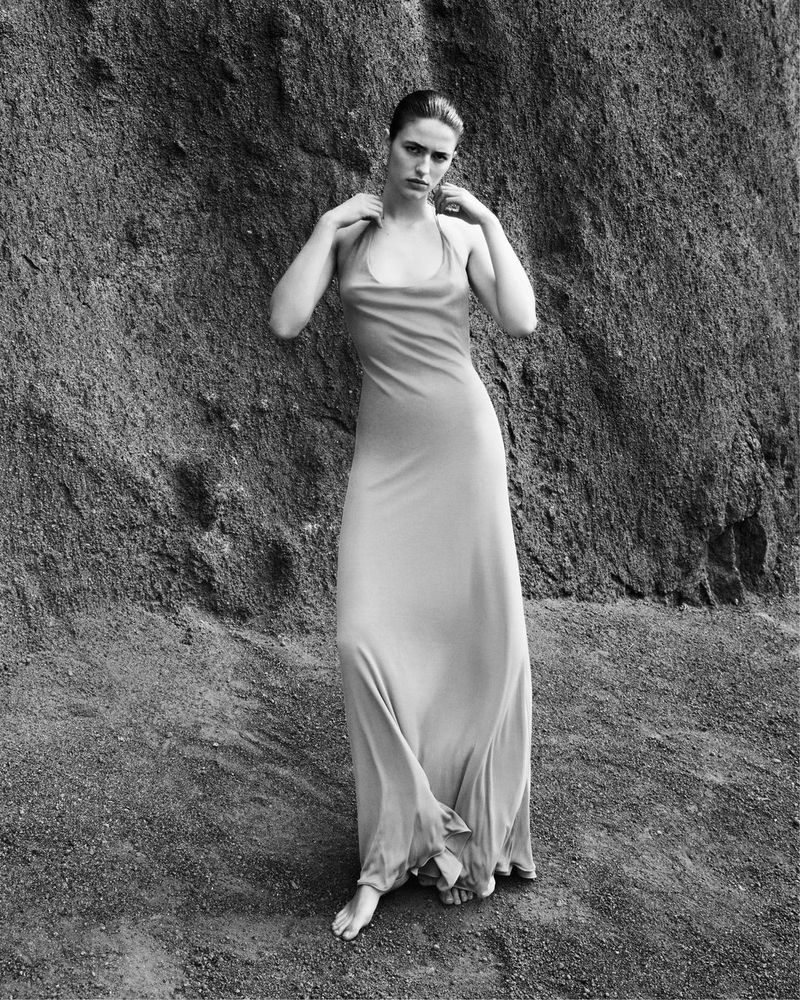 A woman in a light-colored gown stands in front of a large rock formation in an outdoor setting, styled in black and white, with soft lighting.