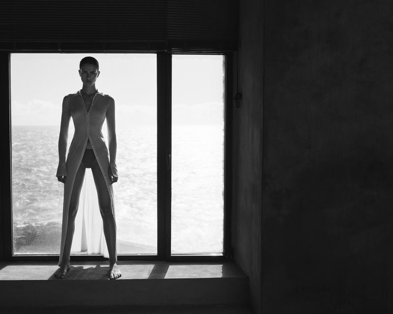 A woman stands near a window looking out at the ocean. The image is black and white with dark wall on one side and bright window.