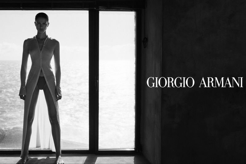 Black-and-white fashion shot of a woman in a translucent robe standing in front of a window, the ocean visible in the background.